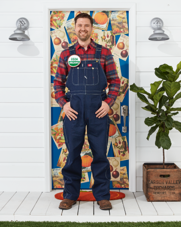 bărbat care poartă costum de fermier cu salopetă, cămașă în carouri, nasture organice usda stând în fața ușii decorate pentru Halloween