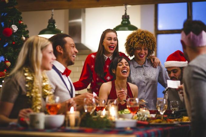 grup de prieteni care râd în timp ce stau în jurul unei mese, mănâncă și beau de Crăciun, un bărbat citește o glumă dintr-un cracker de Crăciun poartă pălării și pulovere de Crăciun