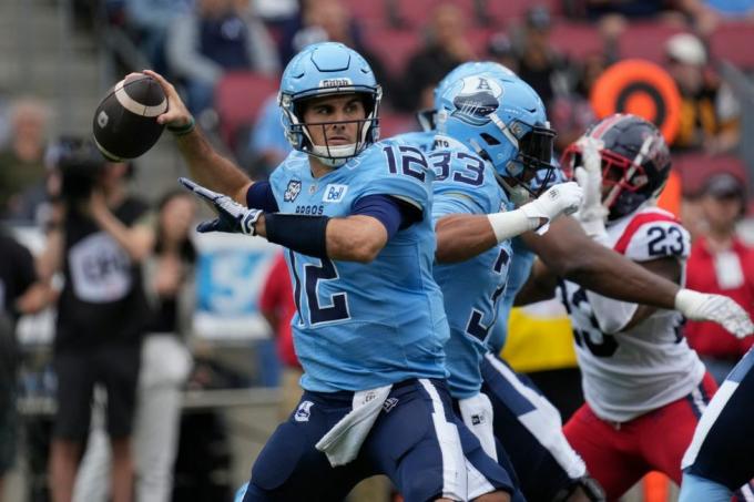 Quarterback de fotbal canadian în uniformă albastră aruncă o pasă în timp ce linia ofensivă reține un jucător defensiv în roșu și alb