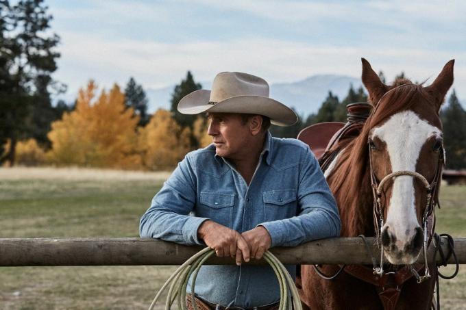 Kevin Costner în Yellowstone lângă un cal sprijinit de un gard cu frânghie în mâini, purtând o cămașă albastră decolorată și pălărie de cowboy bej