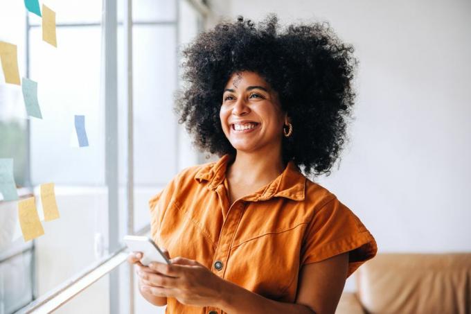 femeie de afaceri neagră fericită zâmbind în timp ce folosește un smartphone într-un birou creativ antreprenor vesel care trimite un mesaj text în timp ce stă lângă un perete de sticlă cu note lipicioase