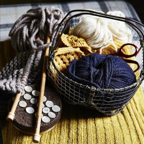 bile de lână albastră și albă în coș mic de sârmă, ace de tricotat din lemn, nasturi, așezate pe țesătură tricotată cu nervuri galbene, textură