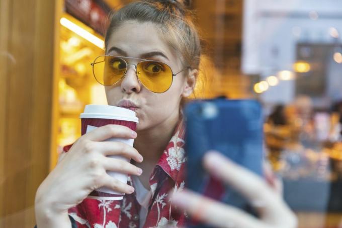 tânără afară, cu ochelari de soare, făcând un selfie în timp ce bea o cafea